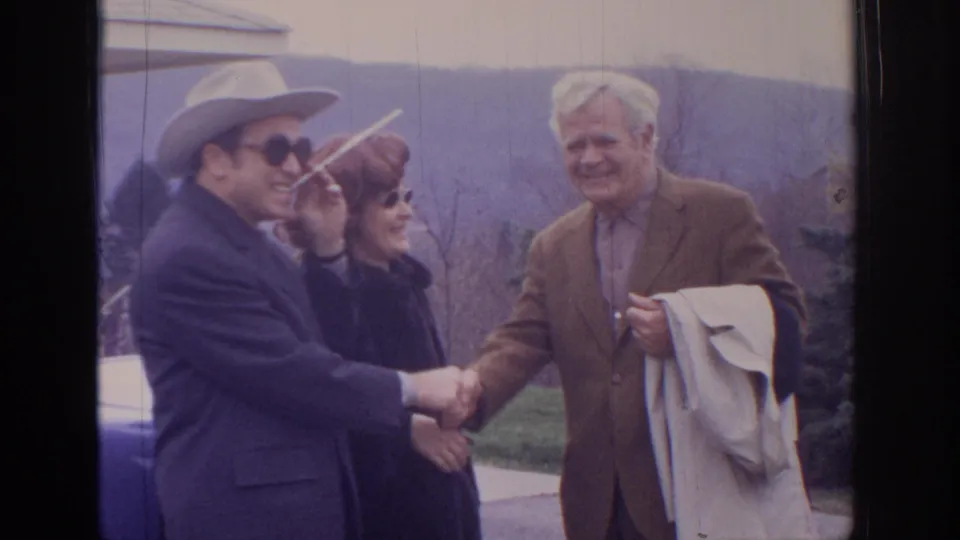1972 home-movie frame of a handshake outdoors, with a woman in sunglasses standing between the two men and hills in the background.