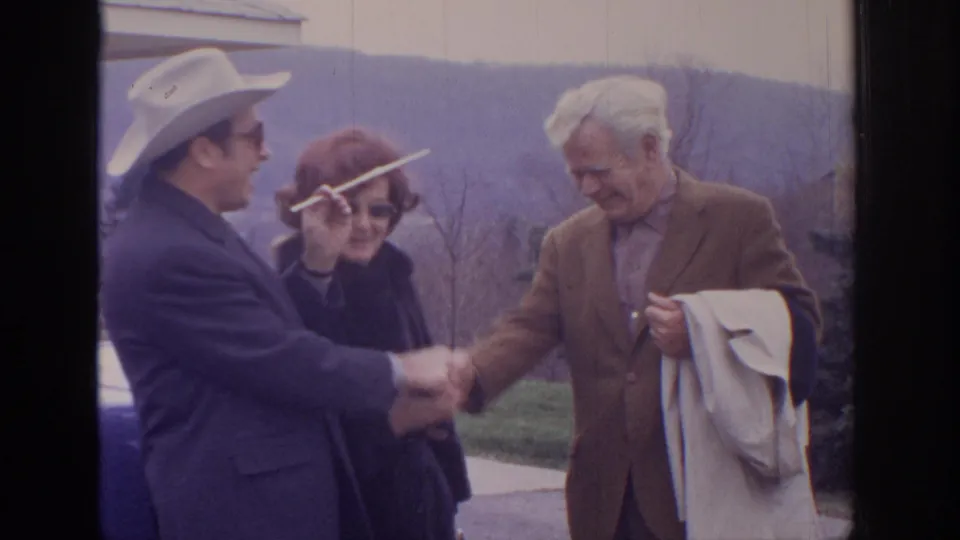 1972 home-movie frame showing a continued handshake between two men outdoors, with a woman watching and wooded hills behind them.