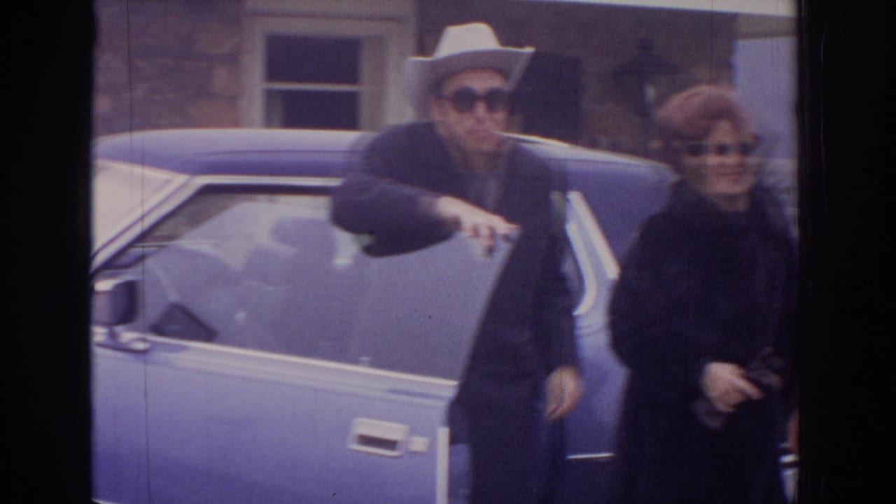 A man in a white cowboy hat and a woman in sunglasses stand beside a blue car.