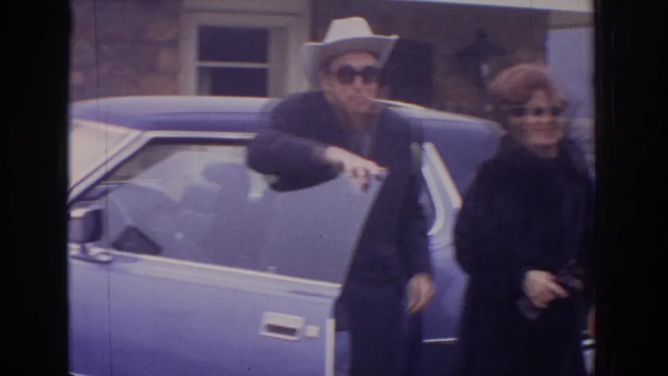 A man in a white cowboy hat and a woman in dark sunglasses stand beside a blue sedan in 1972 Philadelphia home-movie footage.