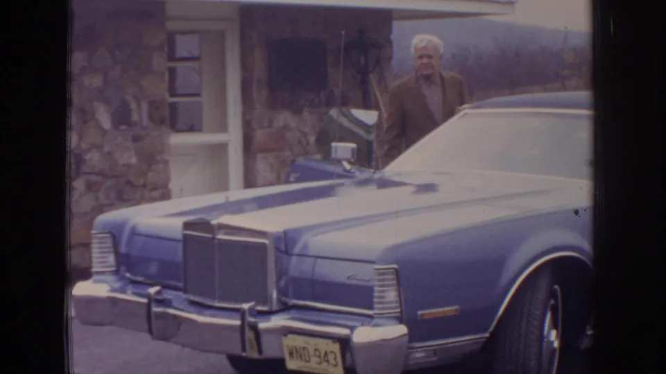 An older man stands near the chrome front end of a blue sedan outside a stone building, with a yellow license plate visible.