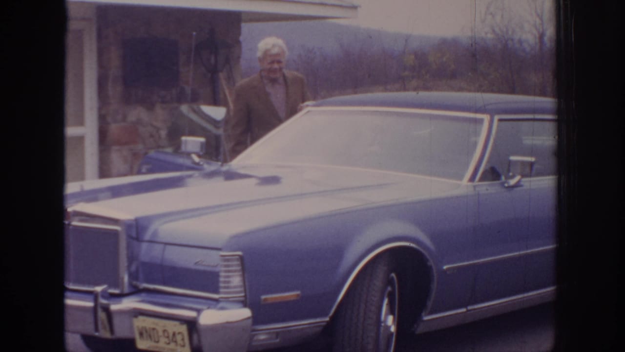 Older man stands behind the front of a blue car near a house exterior.