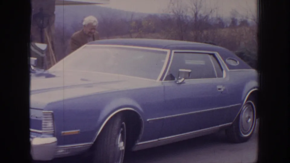 1972 home-movie frame showing a blue classic sedan in a driveway with an older man partly visible behind the roofline.