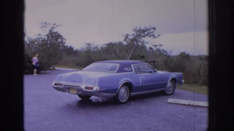 1972 Philadelphia-area 8mm frame showing a blue 1970s coupe parked near brush, with a distant person walking at the edge of the lot.