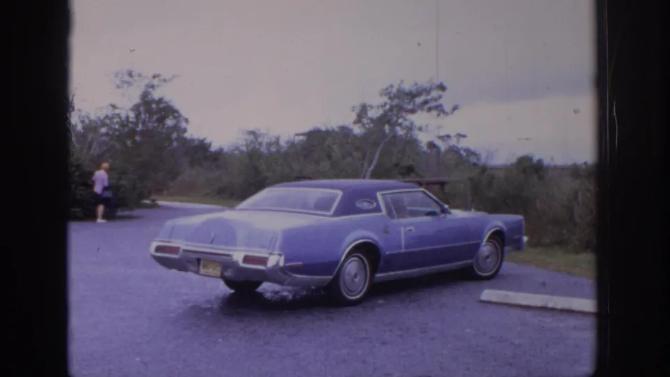 1972 Philadelphia-area 8mm frame of a blue coupe in a paved or gravel lot, with brush behind it and a distant person near the road.