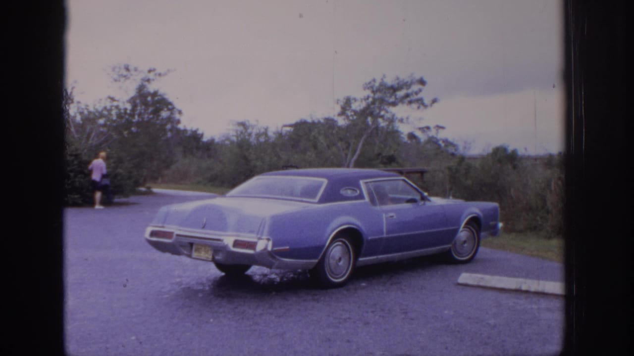 1972 Philadelphia-area 8mm frame showing a blue classic sedan parked on roadside pavement with trees and brush behind it.
