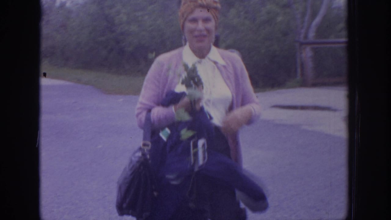Woman in a headscarf and pink cardigan walks toward the camera holding flowers and bags.