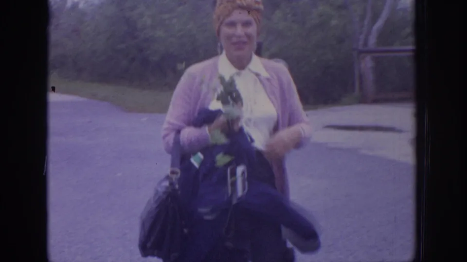 1972 Philadelphia-area 8mm frame of a smiling woman in a headscarf and pink cardigan carrying flowers and belongings outdoors.