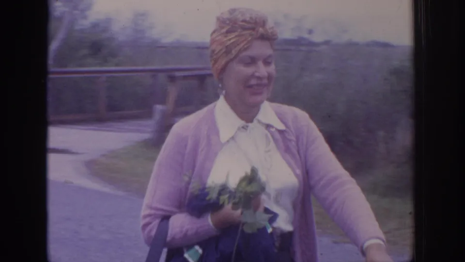 1972 Philadelphia-area home-movie frame showing the woman smiling beside a road with flowers in hand and trees behind her.