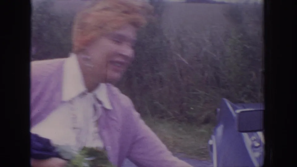 1972 Philadelphia-area 8mm frame showing the woman passing close to the camera near the blue sedan, with motion blur and greenery behind.