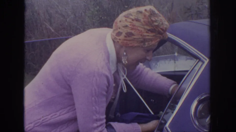 A woman wearing a patterned headscarf and purple sweater leans into the open blue sedan in the stated 1972 home-movie clip.