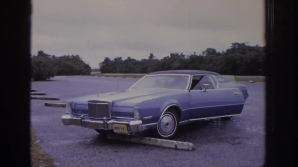 A blue early-1970s sedan sits in a paved parking lot with trees and cloudy sky in the background.