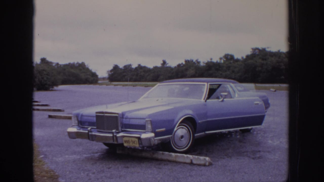 1972 8mm home-movie frame of a blue classic car parked beside a curb stop in a gravel lot, with trees and gray sky behind it.