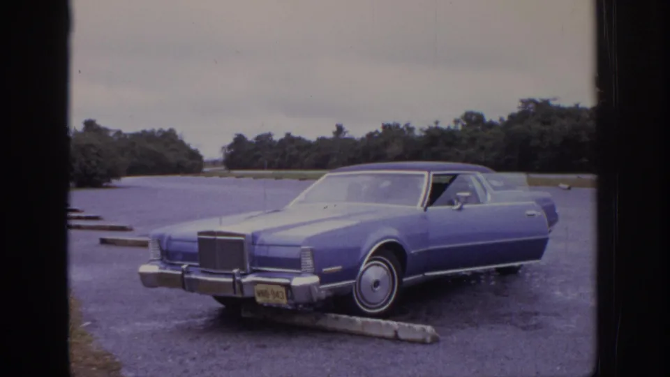 1972 8mm home-movie frame of a blue classic car parked beside a curb stop in a gravel lot, with trees and gray sky behind it.