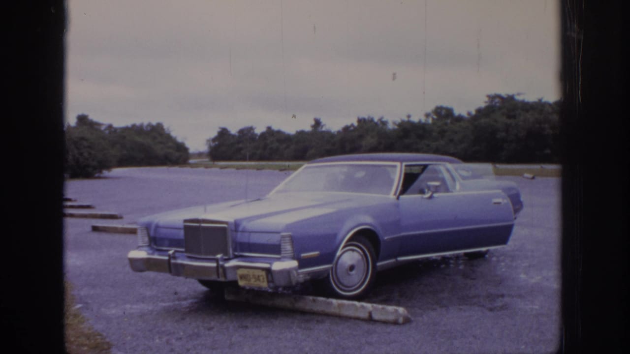 Blue 1970s-era Lincoln-style car remains framed in a gravel lot, with chrome trim, a front license plate, trees, and an overcast sky visible.