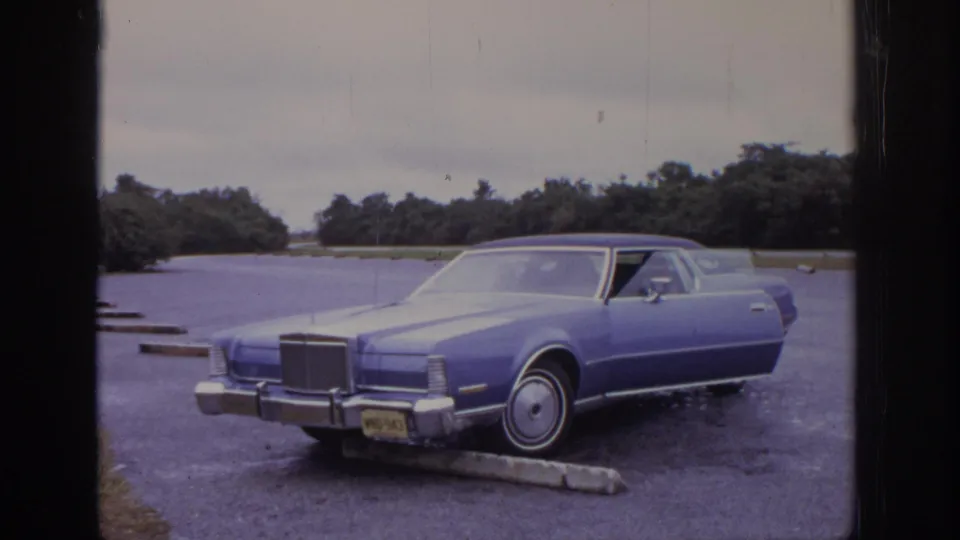 Blue 1970s-era Lincoln-style car remains framed in a gravel lot, with chrome trim, a front license plate, trees, and an overcast sky visible.