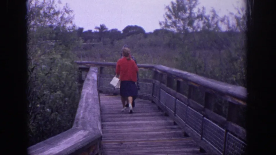 A woman in a red sweater walks away along a wooden boardwalk in the supplied 1972 Philadelphia nature footage.