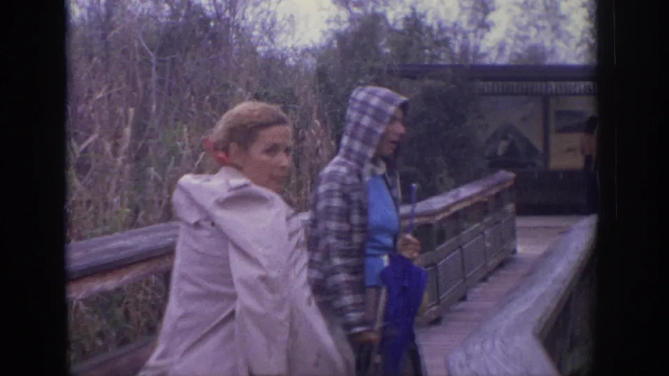 Two women pause near the boardwalk railing; one wears a plaid hooded jacket and holds a blue umbrella in the 1972 Philadelphia footage.
