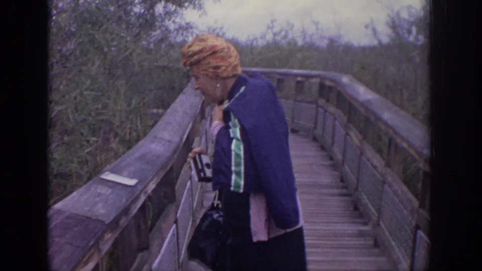 A woman with a patterned headscarf and camera stands beside the wooden railing on the Philadelphia boardwalk in the 1972 home movie.