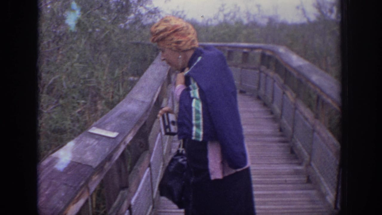 1972 8mm home-movie frame showing a woman in a patterned headscarf and blue jacket pausing on a wooden boardwalk in Philadelphia metadata context.