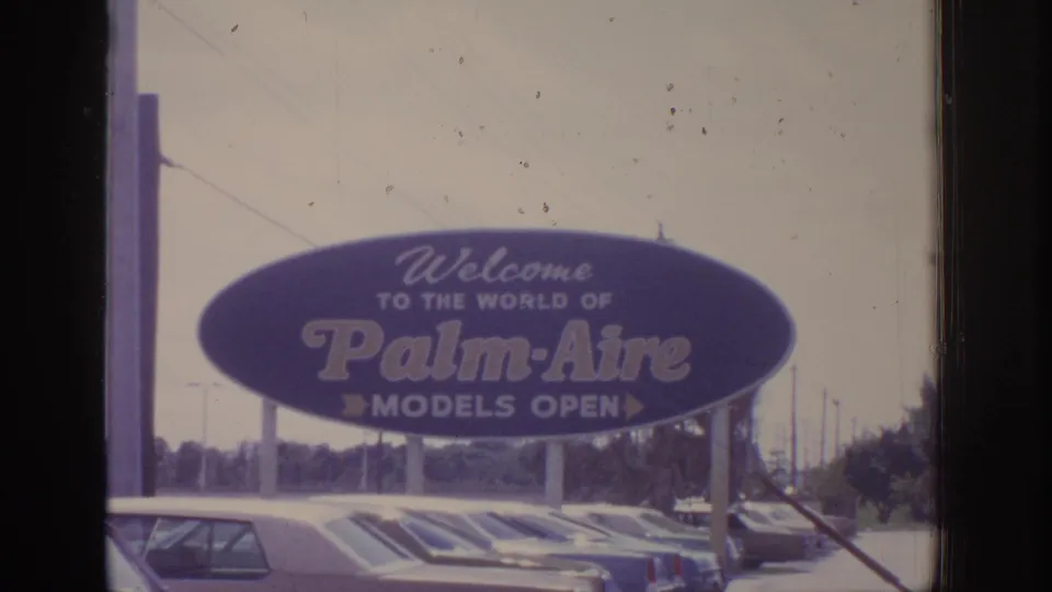 Faded 8mm frame of a Palm-Aire roadside welcome sign and parking area, with legible branding requiring rights review.