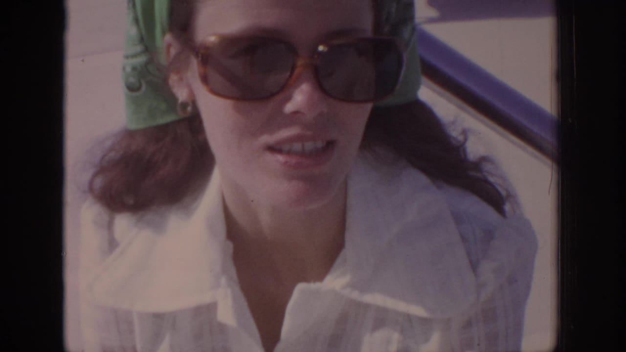Adult woman in 1972 home-movie footage wears sunglasses, a green bandana, and a white collared shirt near a car window.