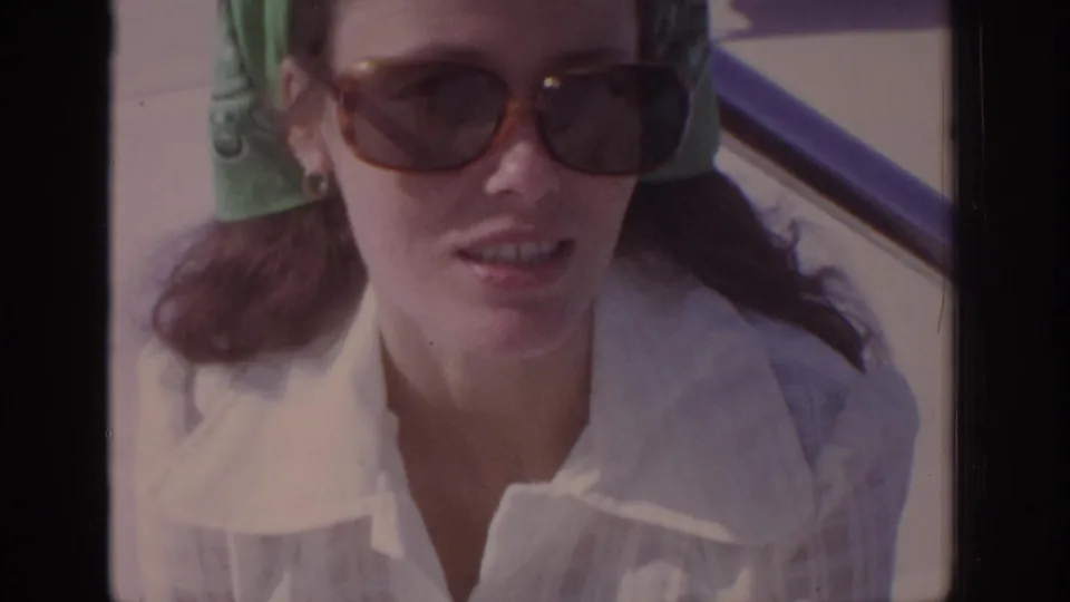 Adult woman in 1972 home-movie footage wears sunglasses, a green bandana, and a white collared shirt near a car window.