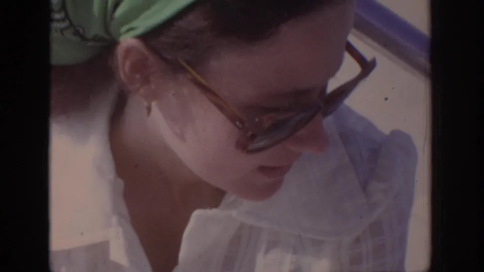 Close 1972 8mm frame of an adult woman with sunglasses, green bandana, and white collared shirt looking down near a car.