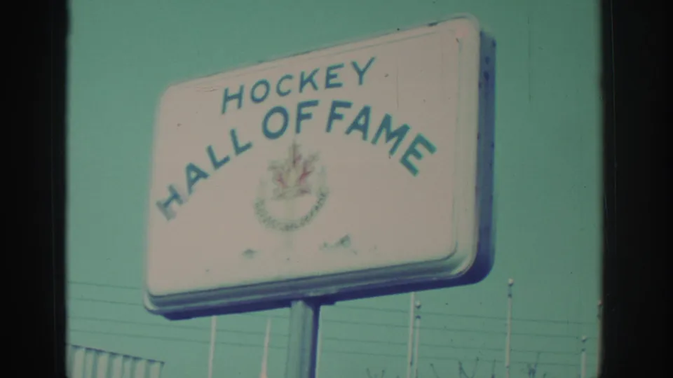 Closer 8mm view of the Hockey Hall of Fame sign, with lettering, emblem, support posts, and flagpoles visible nearby.