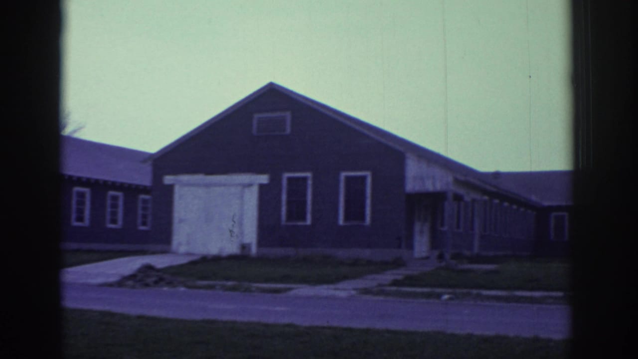 1972 Toronto home-movie frame showing a low dark building, white-trimmed windows, a paved drive, lawn, and pale sky.