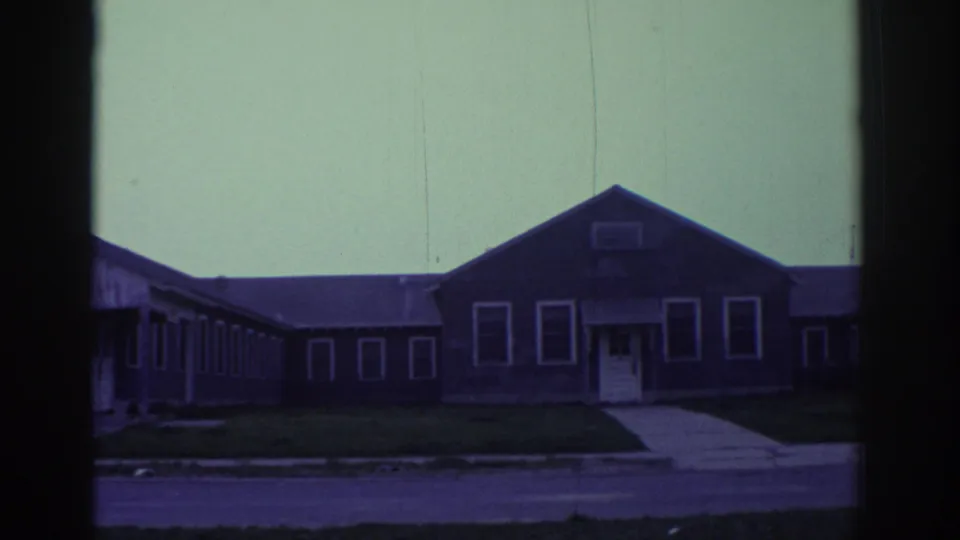 A gabled portion of the 1972 Toronto building appears with white-trimmed windows, an entrance, lawn, sidewalk, and street foreground.