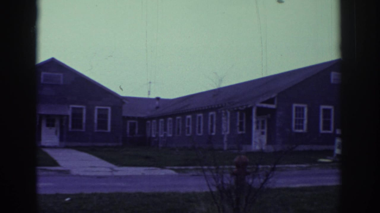 1972 Toronto-linked 8mm frame showing low gray bungalow-style buildings, sidewalks, and dark window-frame edges from a vehicle viewpoint.