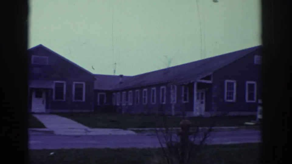 1972 Toronto-linked 8mm frame showing low gray bungalow-style buildings, sidewalks, and dark window-frame edges from a vehicle viewpoint.
