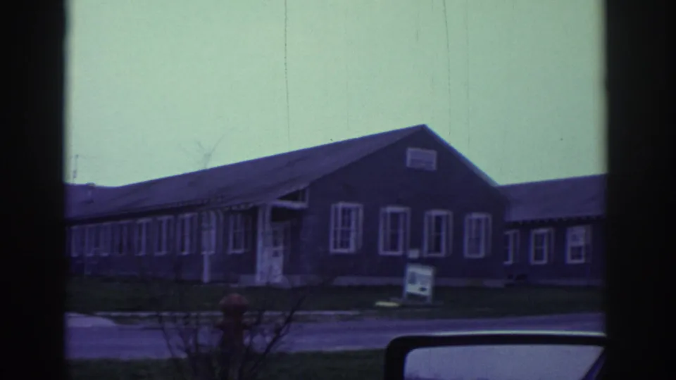 Passenger-window view of gray bungalow-like buildings with a roadside sign, hydrant, and vehicle mirror in 1972 Toronto-linked footage.