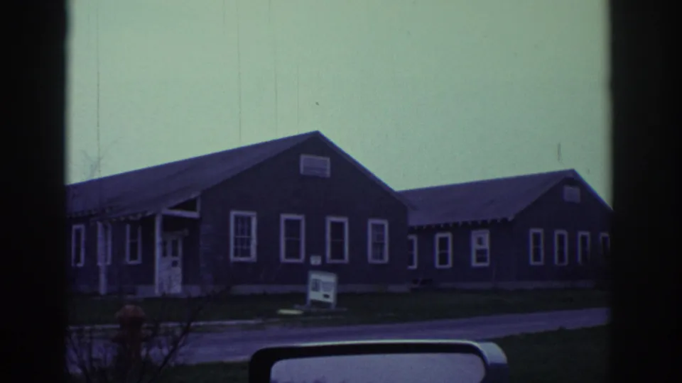 1972 Toronto-linked home-movie frame showing low gray buildings, a small sign, a hydrant, and a vehicle mirror under cloudy light.