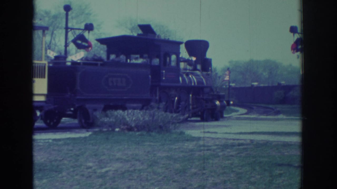 1972 Toronto home-movie frame showing a dark steam-style locomotive and yellow sightseeing car moving through a grassy park setting.