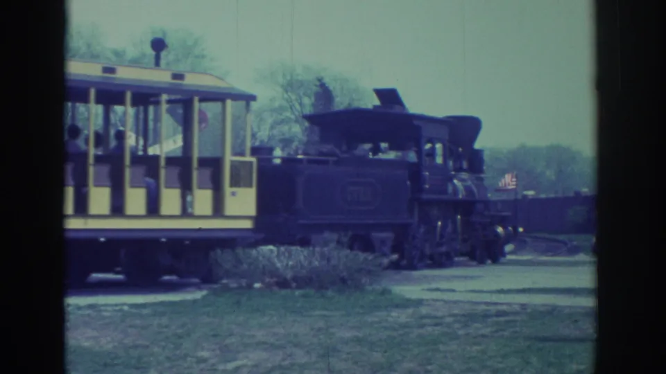 1972 Toronto frame with an open-sided yellow passenger car carrying riders behind a dark steam-style locomotive.