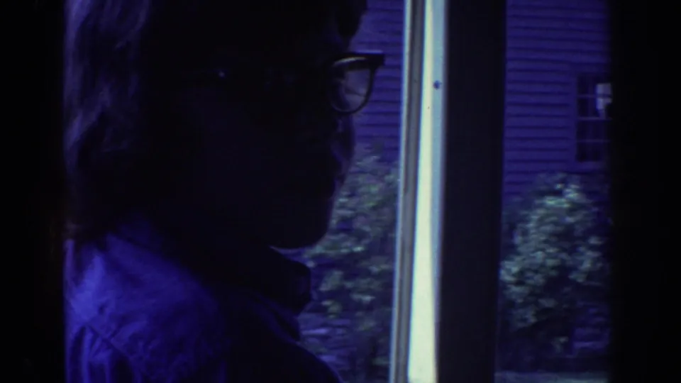 1972 Toronto home-movie frame showing a silhouetted person wearing glasses near a bright vertical window or doorway, with exterior siding beyond.