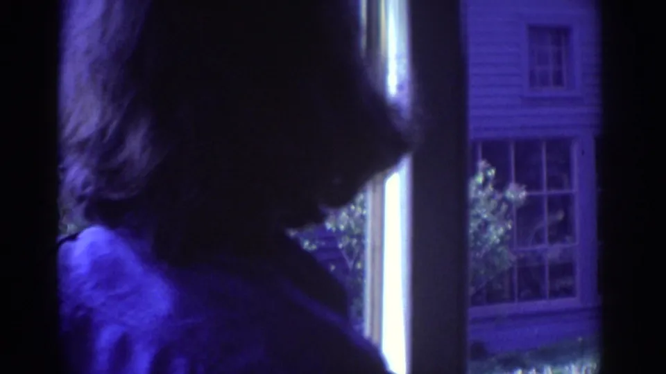 1972 Toronto home-movie frame showing a person from behind near a window, with a clapboard building and multi-pane windows visible outside.