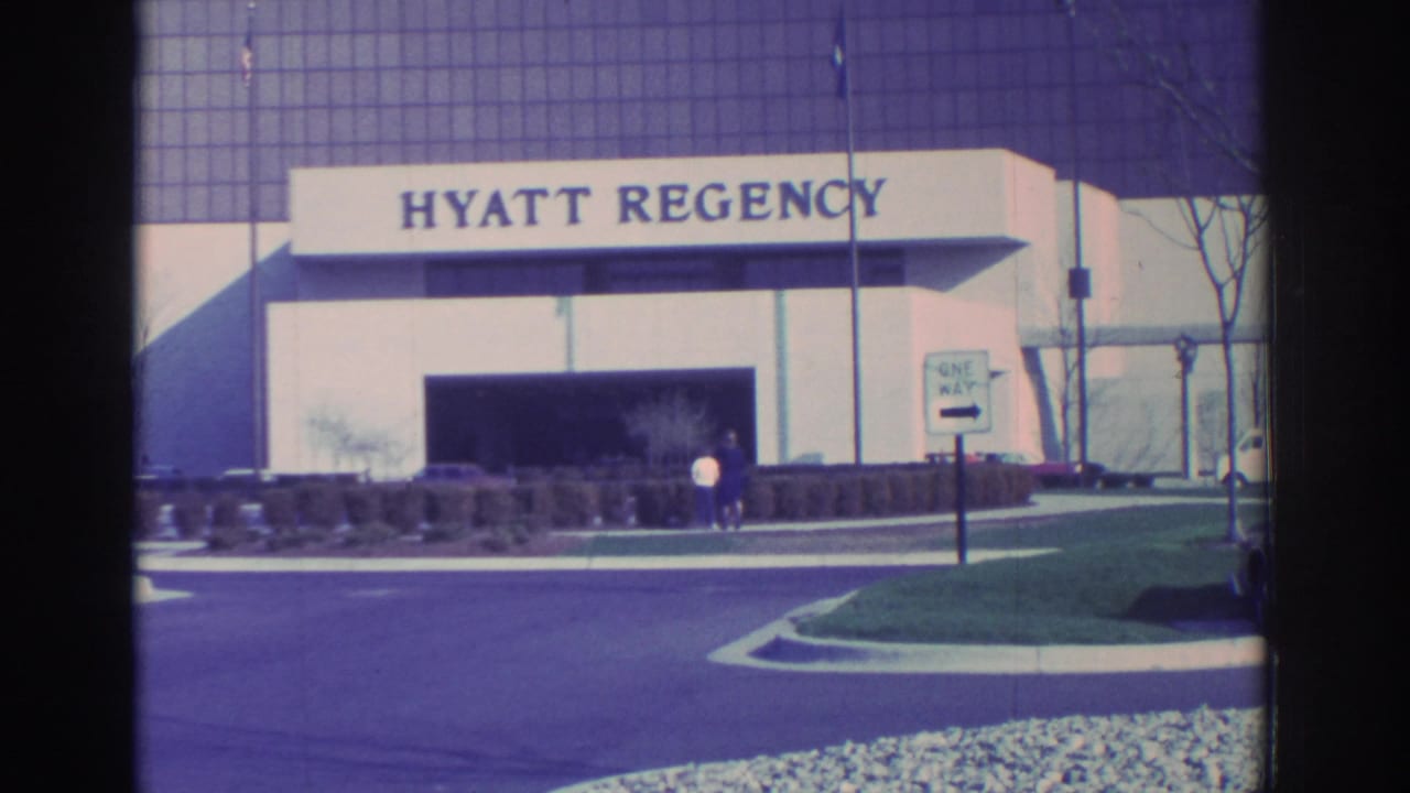 Hyatt Regency entrance with driveway, shrubs, sign, and two people near the doorway.