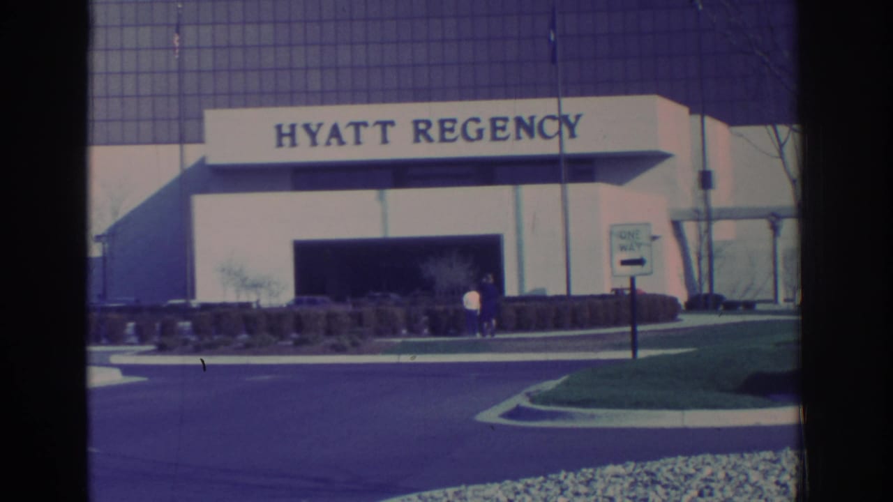 Hyatt Regency entrance and facade with a driveway, sign, shrubs, and two people standing near the doorway.