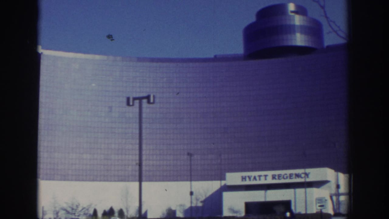 1972 Toronto home-movie view of the Hyatt Regency curved glass facade, rooftop cylinder, blue sky, and streetlight.