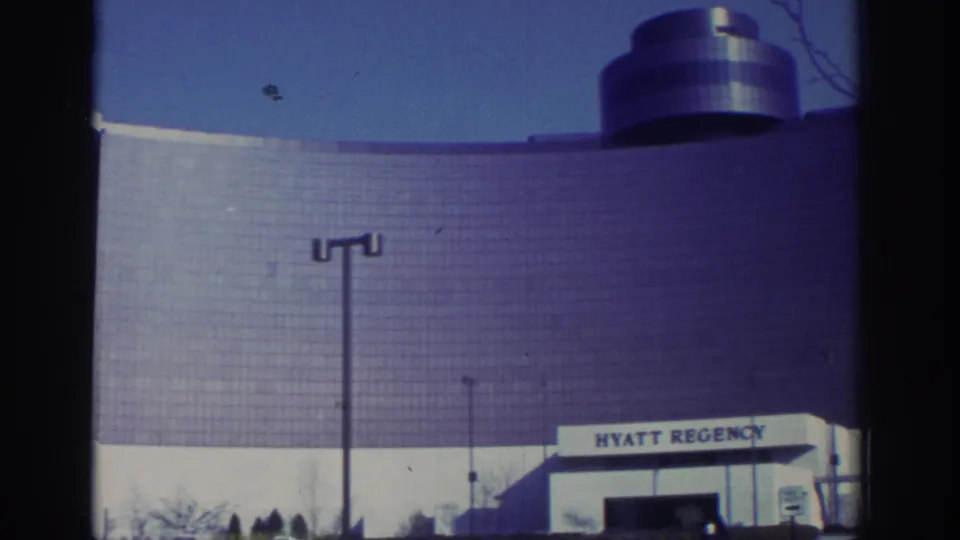 1972 Toronto home-movie view of the Hyatt Regency curved glass facade, rooftop cylinder, blue sky, and streetlight.