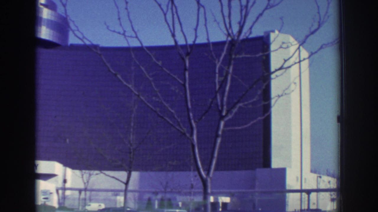 1972 Toronto home-movie frame showing a modern building facade, leafless branches, and part of an elevated transit structure.