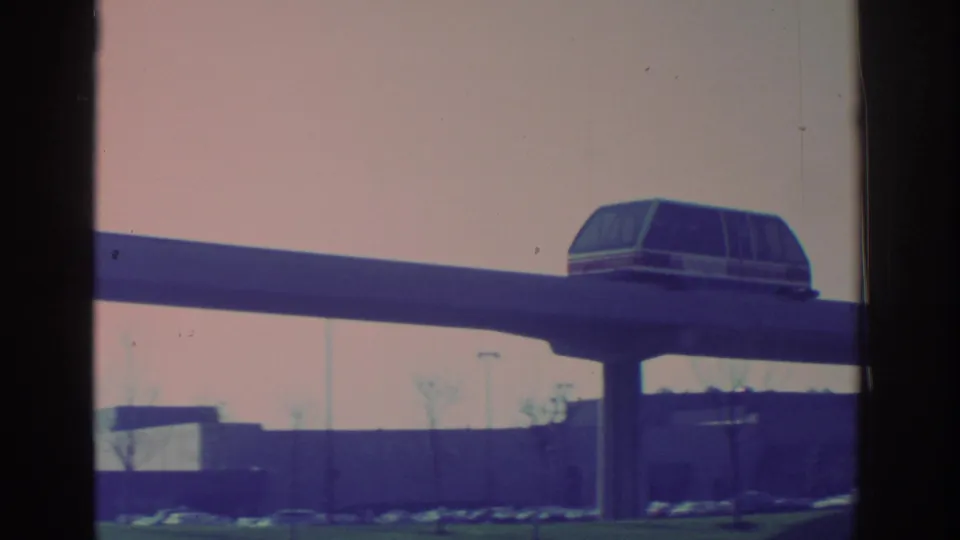 1972 Toronto frame of an enclosed monorail-style car gliding on a raised guideway near low buildings and parked cars.