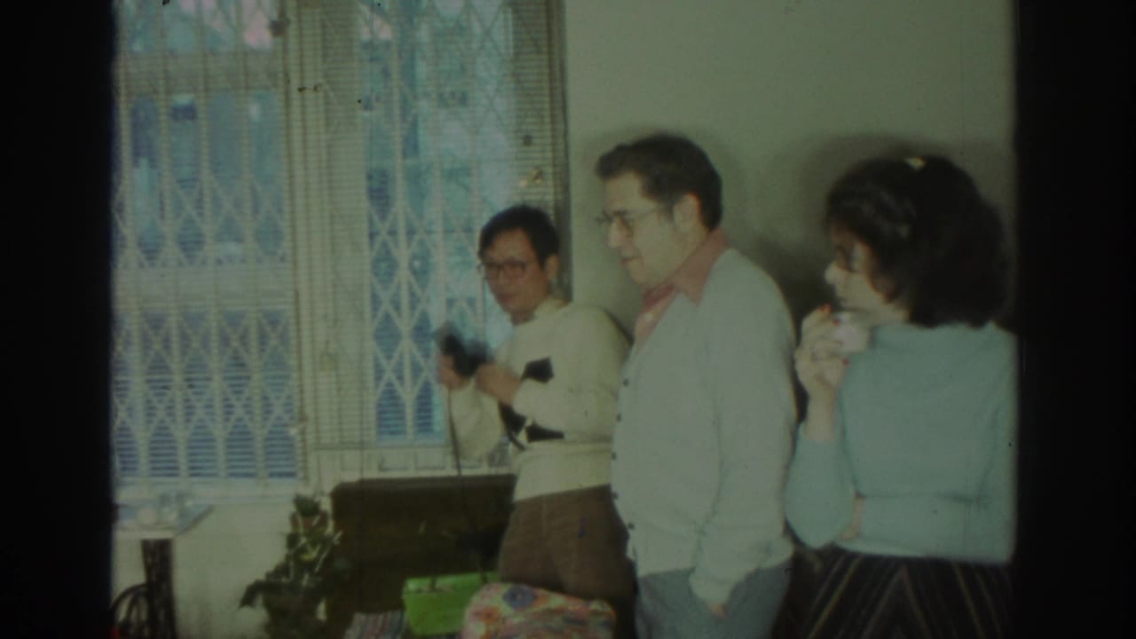 1972 Toronto home-movie frame showing adults in cardigans and sweaters gathered near a curtained window inside a private home.