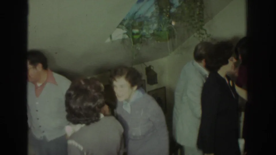 1972 Toronto home-movie frame with adults socializing by a staircase under a hanging plant inside a private residence.
