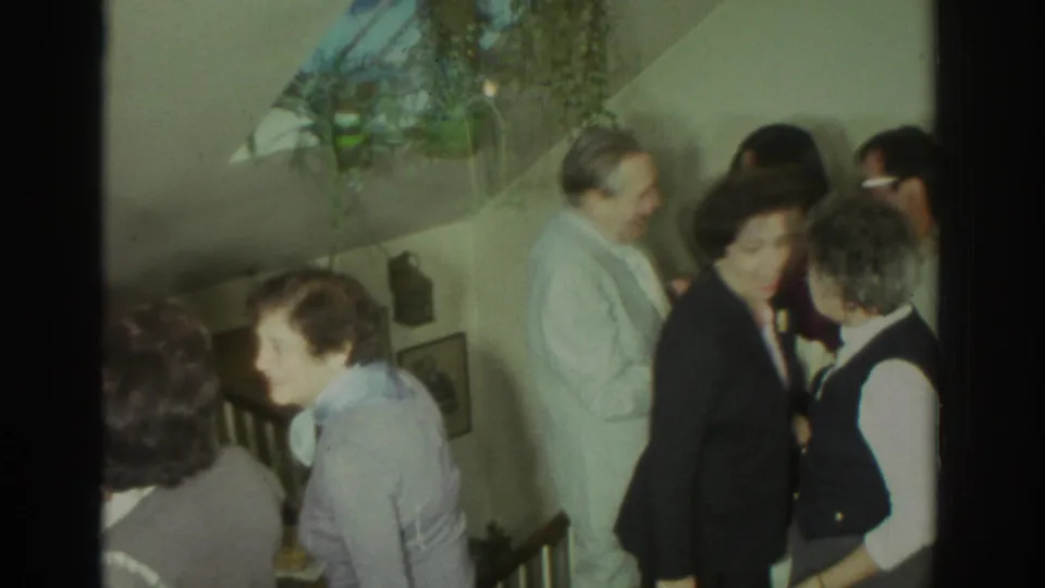 1972 Toronto home-movie frame showing adults in conversation near a staircase, with casual and semi-formal clothing visible.
