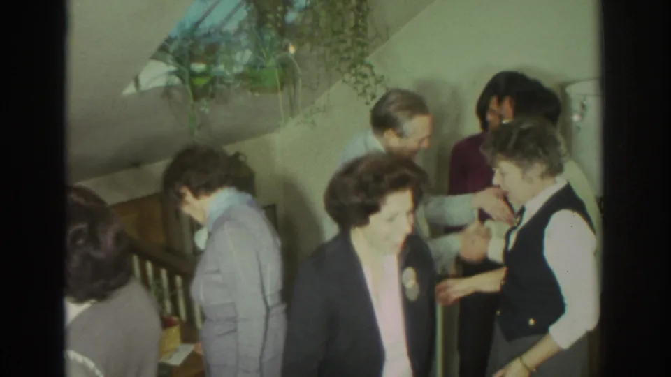 1972 Toronto home-movie frame of adults talking beside a staircase, with a hanging plant and domestic wall decor visible.