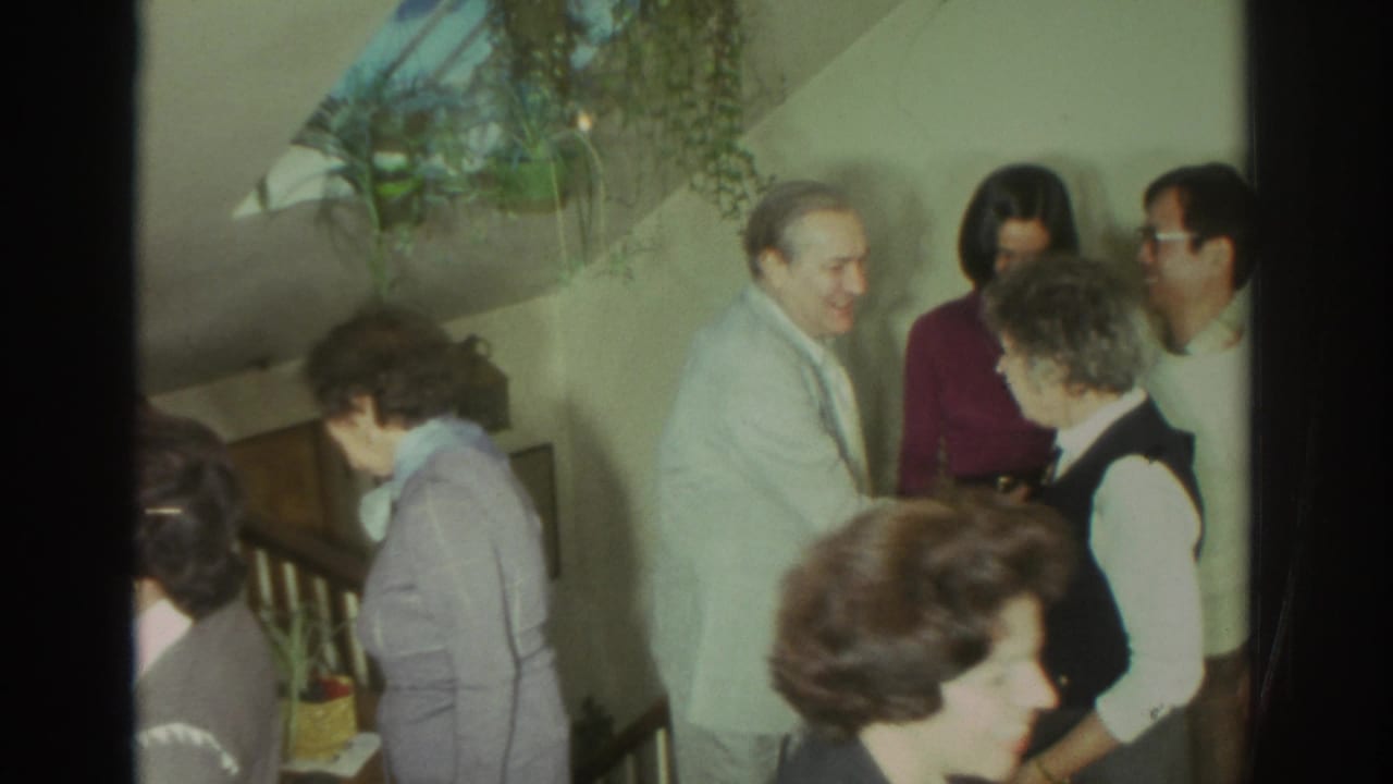1972 Toronto home-movie frame showing adults in a private interior near a staircase, talking and greeting one another.